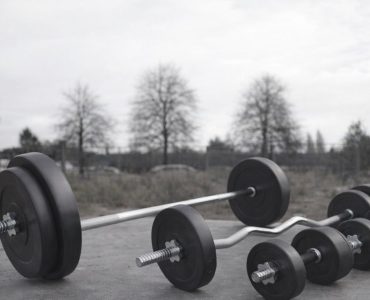 Tre skivstänger och hantlar med viktskivor ligger på marken utomhus.