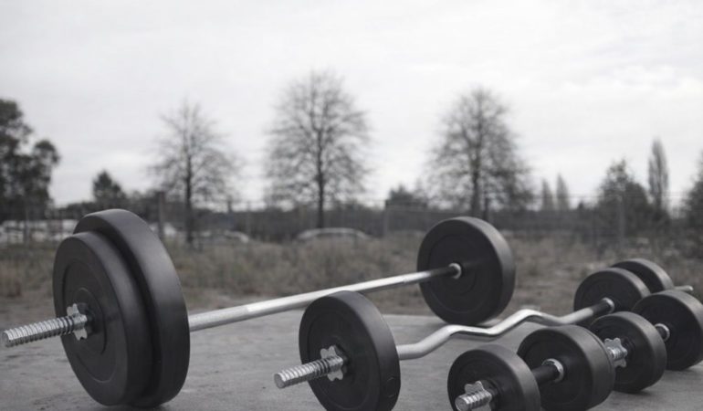 Tre skivstänger och hantlar med viktskivor ligger på marken utomhus.